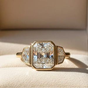 Könnte beinhalten: Ein goldener Ring mit einem großen, rechteckigen, klaren Edelstein als Mittelpunkt, flankiert von zwei kleineren, trapezförmigen Steinen. Der Ring befindet sich in einer braunen Samtbox mit dem goldenen Schriftzug "RARE BIJOU".