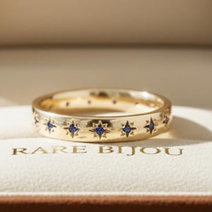 May include: A gold ring with a star pattern and blue gemstones, presented in a brown jewellery box. The box is open, showing the ring on a white cushion. The text "RARE BIJOU" is visible on the box.