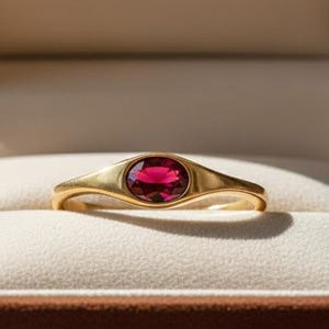 May include: A gold ring with a vibrant red oval gemstone, displayed in a brown jewelry box. The box is open, revealing the ring against a cream-colored interior. The text "RARE BIJOU" is visible inside the box.