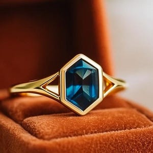 Può includere: Un anello d'oro con una gemma blu scuro esagonale. L'anello è incastonato in una fascia d'oro con un design a gambo diviso. Viene presentato in un portagioie di velluto color ruggine aperto.