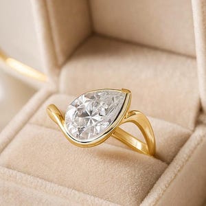 May include: A gold ring featuring a pear-cut diamond in a bezel setting. The ring is presented in a beige velvet box. The band has a distinctive, flowing design. The diamond is highly reflective.