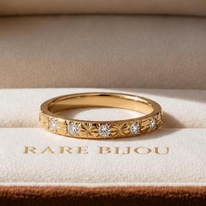 May include: A gold band ring with a repeating starburst and diamond pattern, displayed in a brown velvet jewelry box. The box is open, revealing the ring on a white satin interior. The text "RARE BIJOU" is printed on the box.