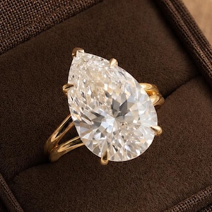 May include: A pear-shaped clear gemstone ring set in a polished yellow gold band. The ring features a large, faceted teardrop-shaped stone held by multiple prongs, resting inside a dark brown, fabric-lined jewellery box with a hinged lid.