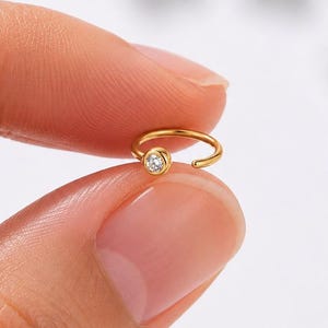May include: A small, gold-colored nose ring with a single, clear, round gemstone. The ring is held between two fingers against a soft, neutral background. The jewelry is delicate and minimalist.