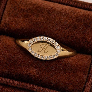 Pode incluir: Um anel de sinete dourado com uma face oval, incrustado com pequenas pedras transparentes. O centro do anel apresenta a letra "K" em uma escrita delicada. O anel é exibido em uma caixa de anel de veludo marrom escuro.