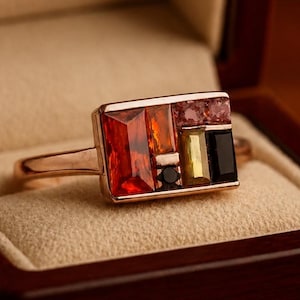 May include: A gold ring featuring a rectangular cluster of multi-colored gemstones, including orange, yellow, black, and pink stones arranged in a geometric pattern, resting inside a dark wooden jewelry box with a beige interior lining.