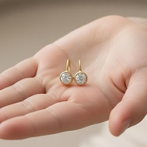 K&ouml;nnte beinhalten: Ein Paar goldfarbene Ohrringe mit einem runden, klaren Edelstein, der in einer L&uuml;nette gefasst ist. Die Ohrringe werden in einer offenen Hand pr&auml;sentiert, um ihr Design hervorzuheben. Die Ohrringe haben ein schlichtes, elegantes Design.
