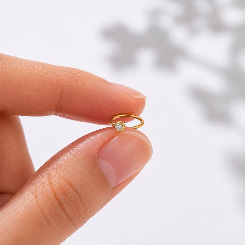 May include: A small, gold-colored nose ring with a single, clear, round gemstone. The ring is held between two fingers against a soft, neutral background. The jewelry is delicate and minimalist.