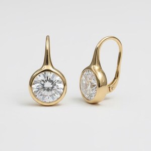 K&ouml;nnte beinhalten: Ein Paar goldene Klappb&uuml;gel-Ohrringe. Jeder Ohrring hat einen runden, klaren Edelstein, der in einer goldenen L&uuml;nette gefasst ist. Die Ohrringe haben ein schlichtes, elegantes Design.