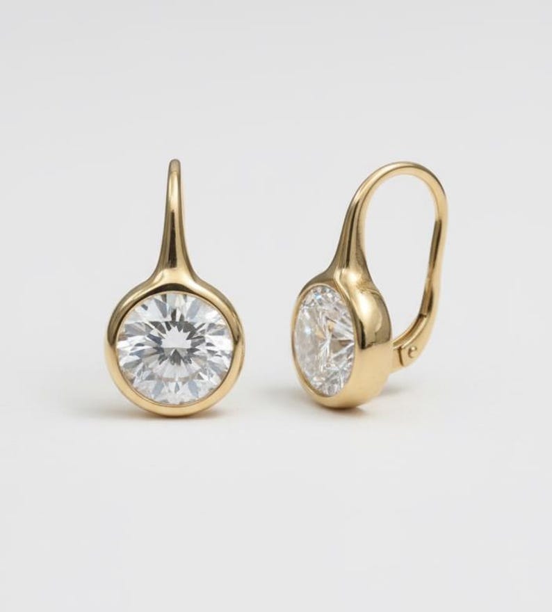 K&ouml;nnte beinhalten: Ein Paar goldene Klappb&uuml;gel-Ohrringe. Jeder Ohrring hat einen runden, klaren Edelstein, der in einer goldenen L&uuml;nette gefasst ist. Die Ohrringe haben ein schlichtes, elegantes Design.