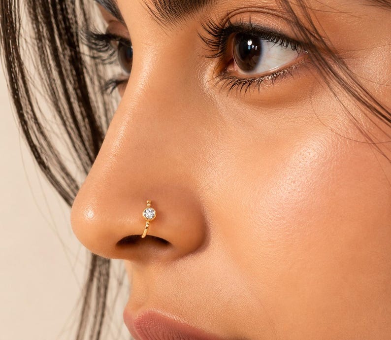 May include: Close-up of a nose with a gold nose ring. The nose ring is a small hoop with a clear, round gemstone at the front. The skin tone is light, and the focus is on the jewelry.