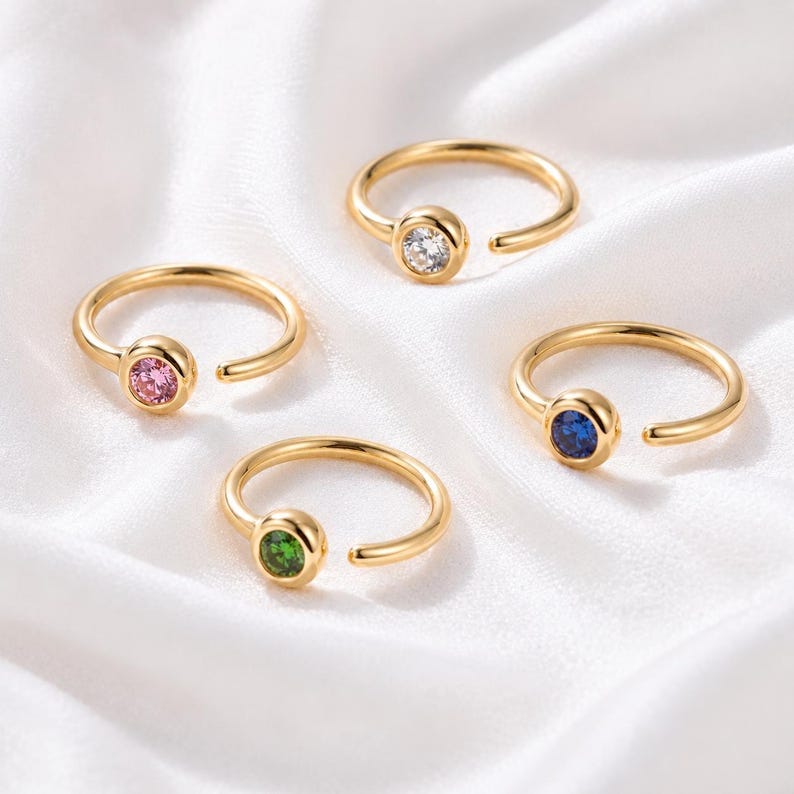 May include: Four gold-colored rings with open bands and bezel-set gemstones. The gemstones are pink, clear, green, and blue. The rings are displayed on a white, silky surface.