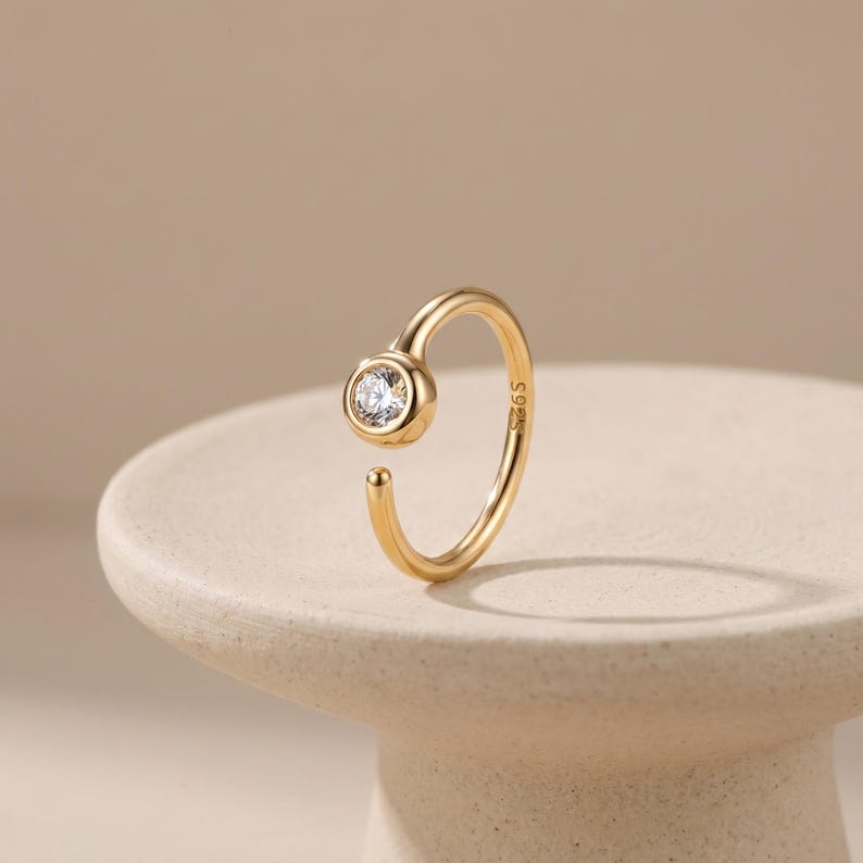 May include: A gold-colored ring with a single, clear gemstone. The ring has an open design, with the gemstone set in a circular bezel. The band curves around to meet the open end. The ring is displayed on a neutral-colored surface.