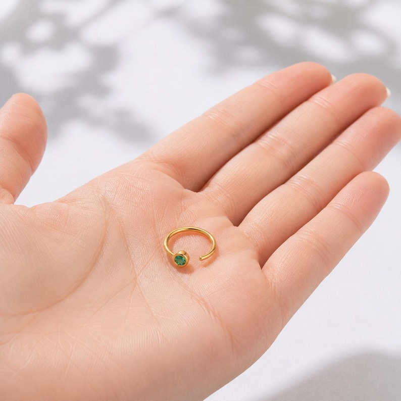 May include: A gold-colored nose ring with a small, round, green gemstone. The ring is open-ended and held in a person's palm. The gemstone is a vibrant green color. The jewelry is simple and elegant.