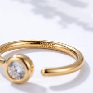 May include: A gold-colored ring with a clear, round gemstone set in a bezel setting. The ring has an open design and the inscription "S925" is visible on the band.