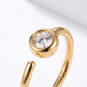 May include: Close-up of a gold-colored ring with a clear, round gemstone. The ring has a curved design with a bezel setting for the gemstone. The metal is stamped with "S925". The ring is likely made of sterling silver.