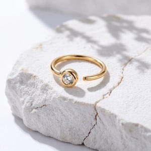 May include: A gold-colored nose ring with a single, clear, round gemstone set in a bezel. The ring is open-ended and rests on a white, textured surface. The jewelry is simple and elegant.