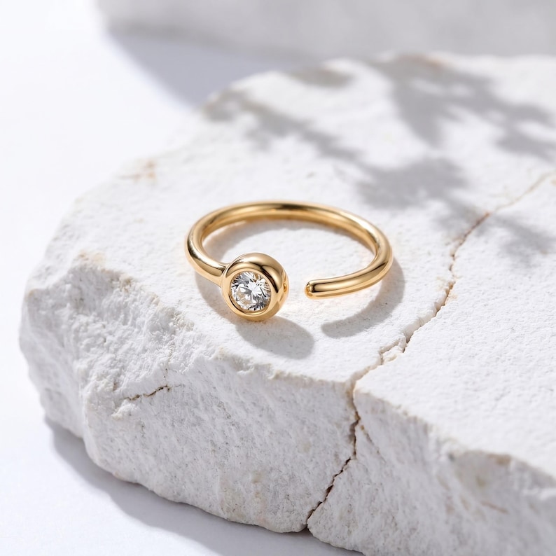 May include: A gold-colored nose ring with a single, clear, round gemstone set in a bezel. The ring is open-ended and rests on a white, textured surface. The jewelry is simple and elegant.