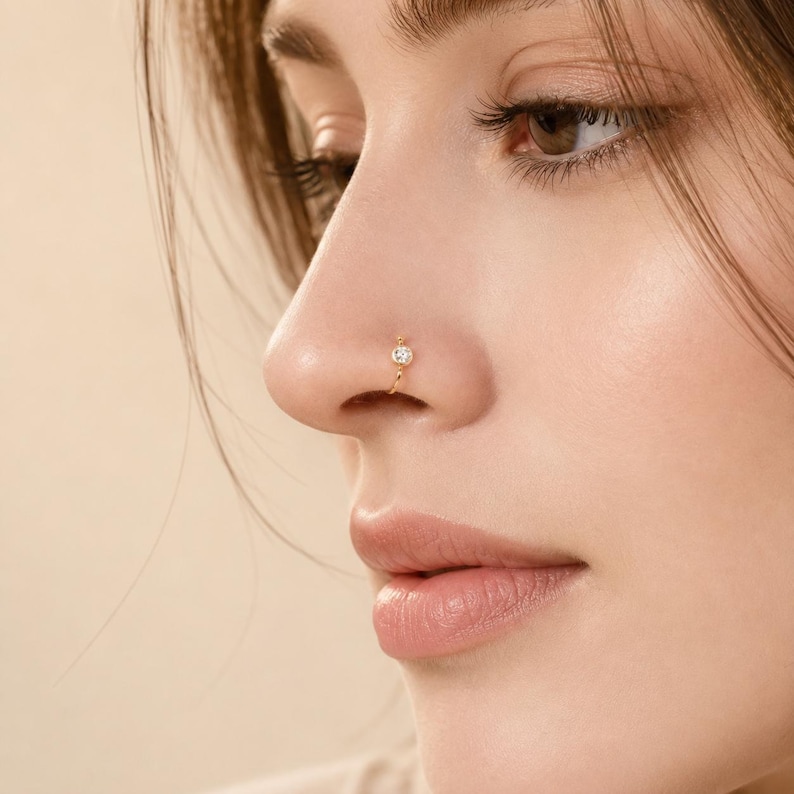 May include: A gold-colored nose ring with a small, clear gemstone at the center. The ring is a simple hoop design, and the gemstone is round. The jewelry is worn on the nose.