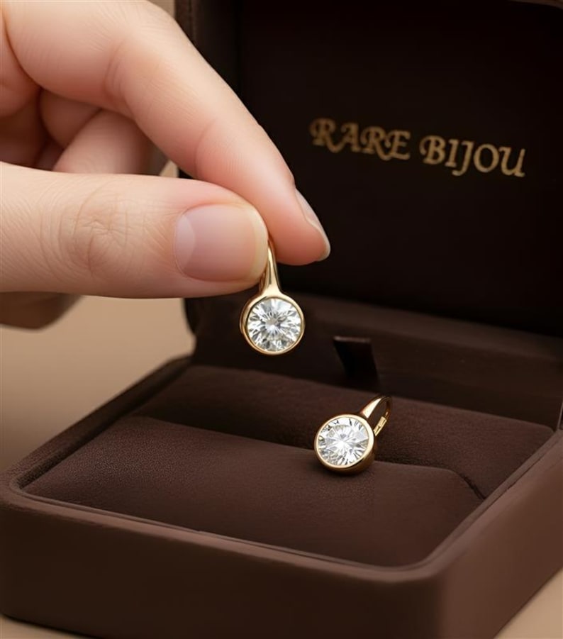 K&ouml;nnte beinhalten: Ein Paar goldfarbene Ohrringe mit runden, klaren Edelsteinen. Ein Ohrring wird von einer Hand gehalten, der andere liegt in einer braunen Schmuckschatulle. Die Schatulle ist ge&ouml;ffnet und zeigt die Ohrringe. Der Text "RARE BIJOU" ist sichtbar.
