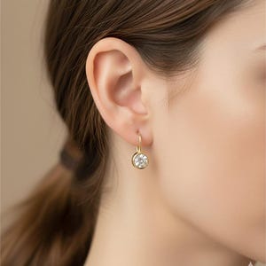 K&ouml;nnte beinhalten: Nahaufnahme eines goldenen Ohrrings mit einem runden, klaren Edelstein. Der Ohrring hat ein schlichtes, elegantes Design mit einem Hakenverschluss. Der Edelstein ist in einer goldenen L&uuml;nette gefasst.