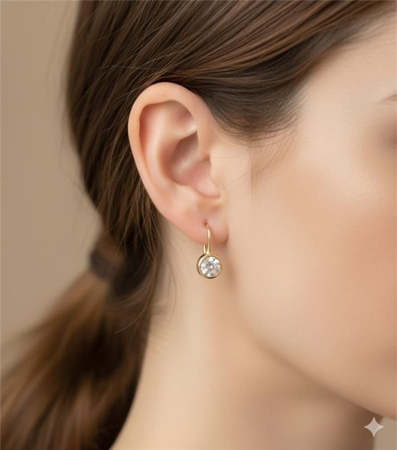 K&ouml;nnte beinhalten: Nahaufnahme eines goldenen Ohrrings mit einem runden, klaren Edelstein. Der Ohrring hat ein schlichtes, elegantes Design mit einem Hakenverschluss. Der Edelstein ist in einer goldenen L&uuml;nette gefasst.