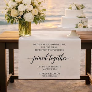 May include: A wedding scene on a beach at sunset. A wooden table holds a floral arrangement in a glass vase, a two-tiered white cake, and a white table runner with text. The text on the runner reads "joined together" and includes names and a date.