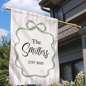 May include: A personalised house flag with a pink and white striped background. The flag features a decorative frame with a bow at the top, and the name "The Smillers" with "EST 2025" in black script.