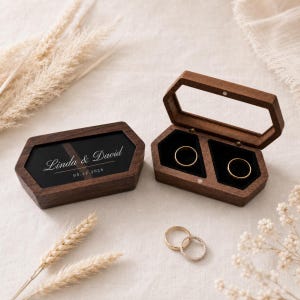 Puede incluir: Dos cajas hexagonales de madera para anillos con marcos de madera oscura y tapas transparentes. Una caja está cerrada, mostrando el texto grabado en blanco Linda & David 05.12.2026. La otra caja está abierta, revelando dos ranuras de terciopelo.