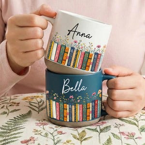 Könnte beinhalten: Zwei Keramikbecher, einer weiß und einer blaugrün, sind gestapelt. Der weiße Becher trägt den Namen "Anna" in schwarzer Schrift. Beide Becher zeigen ein Design aus bunten Büchern und Blumen. Der blaugrüne Becher trägt den Namen "Bella" in Schrift.