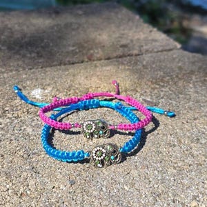 May include: Two handmade friendship bracelets. One is blue, the other is pink, and both have an elephant charm. The elephant charms are silver-colored with green and white accents. The bracelets are on a gray stone surface.