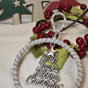 May include: A silver Christmas bauble with a spiral design and a bow. The bauble has a charm that says "Wish You a Merry Christmas" and a star. Wooden Christmas trees and red berries are in the background.