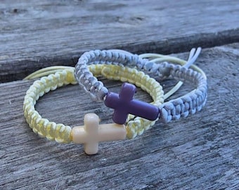 May include: Two braided bracelets with cross charms. One bracelet is light yellow, the other is gray. The cross charms are cream and purple. The bracelets are on a weathered wooden surface.