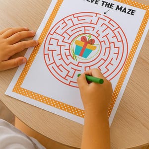 May include: A child's hand holding a green crayon, solving a maze printed on white paper. The maze is red, with a gift box at the center. The paper has an orange dotted border and the text "SOLVE THE MAZE".