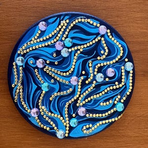 Può includere: Un sottobicchiere rotondo decorativo blu scuro con un design a spirale in tonalità più chiare di blu. Il design è accentuato da punti color oro e abbellimenti iridescenti color gioiello nelle tonalità del viola e del blu.