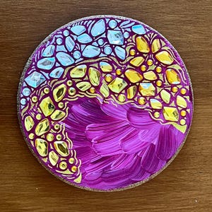 Può includere: Un sottobicchiere rotondo e decorativo con un design vibrante. Il sottobicchiere presenta una combinazione di pennellate viola, accenti dorati e gemme chiare e gialle incastonate. Il bordo esterno ha un bordo strutturato color beige.