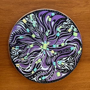 May include: Round coaster with a hand-painted abstract design. The coaster features a central floral-like pattern in shades of purple, black, and white, accented with small, colorful square shapes. The coaster has a dark brown edge.