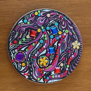 Può includere: Un elemento decorativo rotondo in legno con sfondo nero, adornato con una varietà di gemme colorate e motivi a spirale viola e bianchi. Il design include dettagli rossi, blu, gialli e verdi.