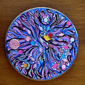 May include: A round, hand-painted coaster with a vibrant, abstract design. The coaster features a blue background with swirling patterns in shades of purple, black, and white. Embellished with colorful gems and glitter accents, it has a cork base.