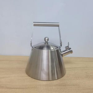 Könnte beinhalten: Edelstahl-Herdkessel mit konischem Körper, klappbarem Griff und Ausguss mit Hebel. Der Kessel hat einen kleinen, runden Knopf auf dem Deckel. Der Kessel steht auf einer Holzoberfläche.