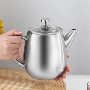 Op de afbeelding: Een roestvrijstalen theepot met een doorzichtig glazen deksel en een kleine, ronde handgreep. De theepot heeft een gebogen tuit en een grote handgreep om gemakkelijk te schenken. De theepot wordt door een persoon vastgehouden.