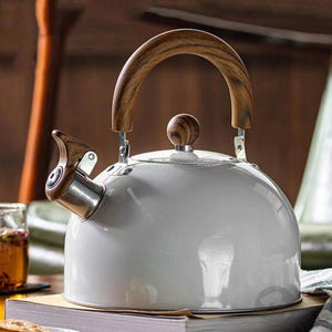 May include: A white enamel tea kettle with a wooden handle and spout. The kettle has a rounded shape and a wooden knob on the lid. The spout and handle are made of wood. The kettle is sitting on a table.