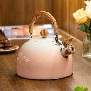 May include: A pink and white ombre tea kettle with a wooden handle and spout. The kettle has a round, bulbous shape and a wooden knob on the lid. The spout also has a wooden accent. The kettle is sitting on a wooden surface.