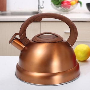 Puede incluir: Una tetera de cocina de color cobre con asa y tapa de madera. La tetera tiene un cuerpo redondeado y un pico. Una fruta amarilla está al lado de la tetera. La tetera está diseñada para hervir agua para té u otras bebidas calientes.