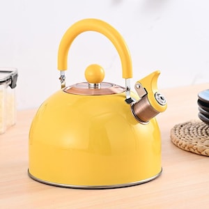 Puede incluir: Una tetera amarilla con un asa curva y un pico. La tetera tiene un cuerpo redondo y un pequeño pomo en la tapa. El asa y el pico también son amarillos. La tetera es de metal y tiene un acabado brillante.