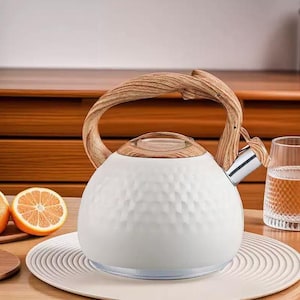 May include: White tea kettle with a textured surface and a wooden handle. The kettle has a stainless steel spout and a clear glass lid. The handle and lid are made of light-coloured wood. The kettle is sitting on a white plate.