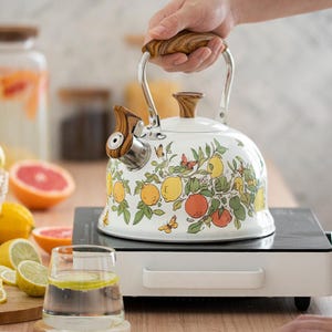 May include: White enamel tea kettle with a fruit and butterfly design. The kettle has a wooden handle and spout, and sits on a white and black electric burner. Lemons and grapefruit are in the background.
