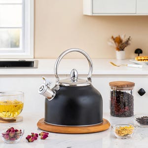 May include: A matte black stainless steel tea kettle with a polished silver handle, lid knob, and spout. The kettle features a whistle mechanism on the spout and sits on a round wooden base, showcasing a sleek, rounded design for stovetop use.