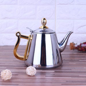 Op de afbeelding: Een gepolijste roestvrijstalen theepot met goudkleurige handgreep en dekseldetails. De theepot heeft een gebogen tuit en een reflecterend oppervlak. De handgreep heeft een kenmerkend, open ontwerp. De theepot staat op een houten oppervlak.