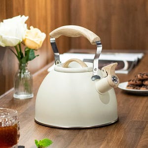 May include: Cream-colored stovetop kettle with a wooden handle and spout detail. The kettle has a stainless steel frame and a wooden knob on the lid. The kettle is sitting on a wooden surface.